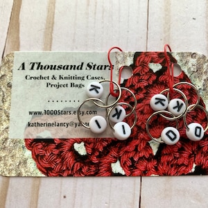 May include: Knitting stitch markers with white letter beads spelling "K I D O" and "K K". The markers are attached to silver rings with red hooks. The background includes a business card with the text "A Thousand Stars".