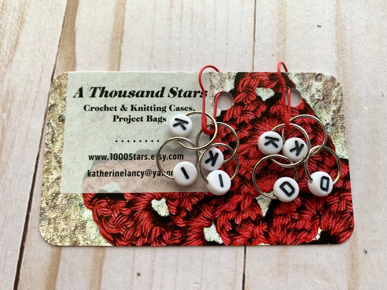 May include: Knitting stitch markers with white letter beads spelling "K I D O" and "K K". The markers are attached to silver rings with red hooks. The background includes a business card with the text "A Thousand Stars".