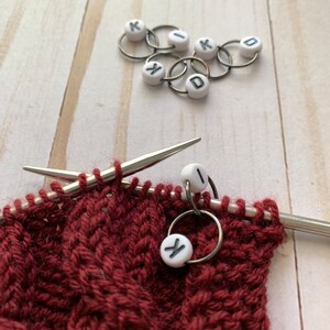 May include: Close-up of knitting project with burgundy yarn, silver needles, and stitch markers. White stitch markers with black letters spell out "KID". The project is in progress, with a cable knit pattern.
