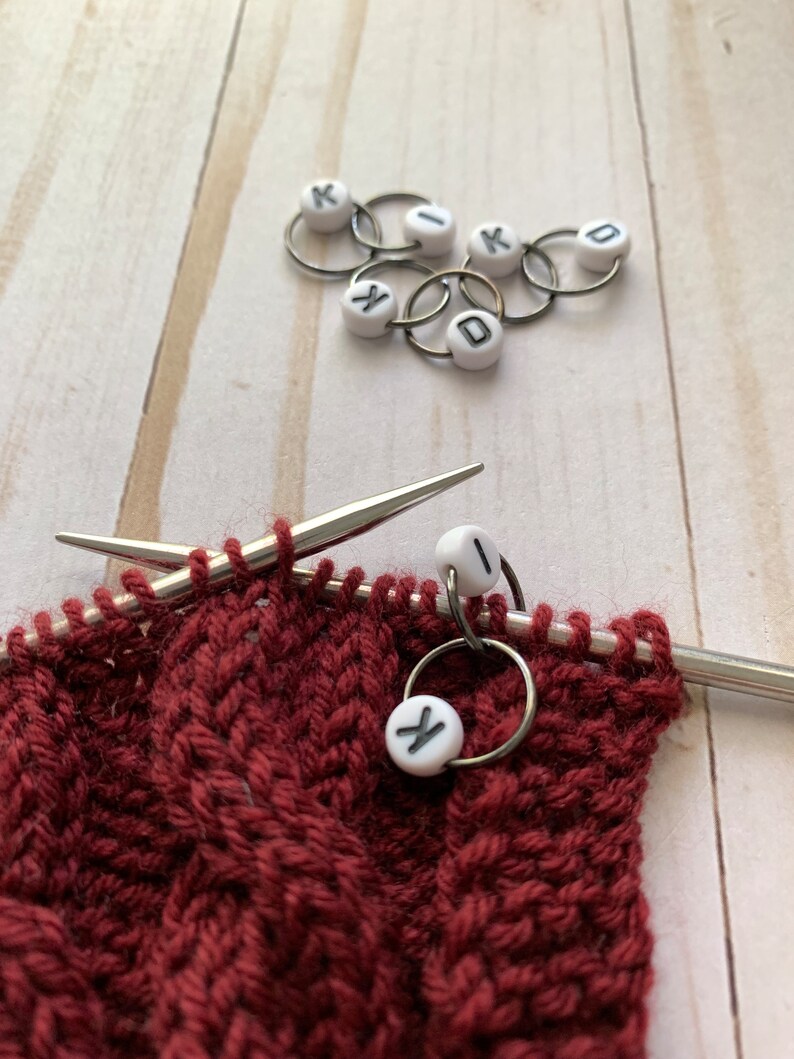 May include: Close-up of knitting project with burgundy yarn, silver needles, and stitch markers. White stitch markers with black letters spell out "KID". The project is in progress, with a cable knit pattern.