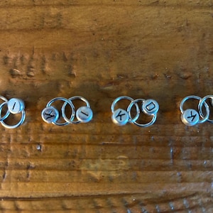 May include: Four silver hoop earrings with small, round, silver beads. Each bead has a single black letter: A, I, K, and D. The earrings are displayed on a wooden surface.
