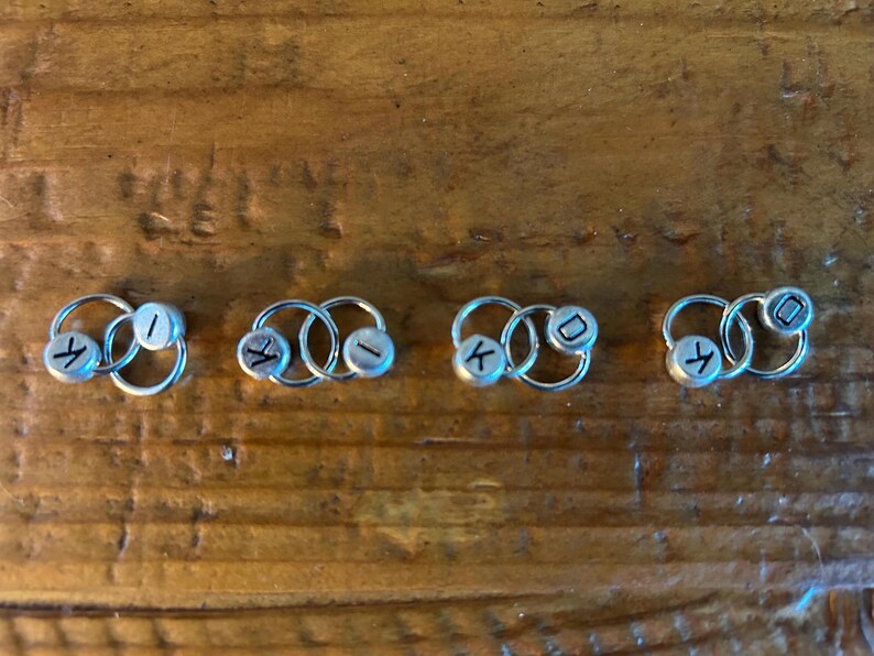 May include: Four silver hoop earrings with small, round, silver beads. Each bead has a single black letter: A, I, K, and D. The earrings are displayed on a wooden surface.