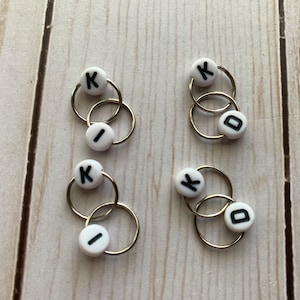 May include: Four sets of small, silver-colored hoop earrings. Each set has two interlocking hoops and two white beads with black letters spelling out "KID". The earrings are displayed on a light wood-grain surface.