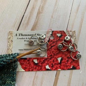 May include: A Thousand Stars Crochet & Knitting Projects business card with a red crocheted sample and silver letter markers with the letters K, I, D, and Y.