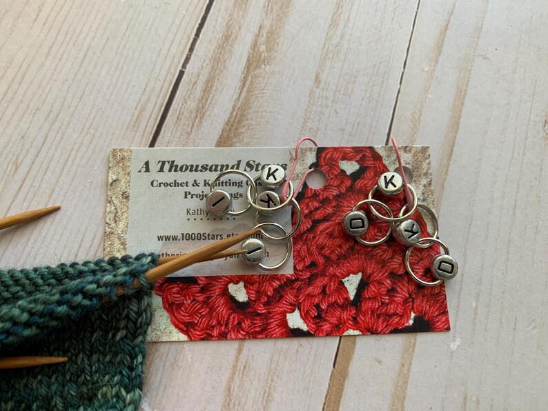 May include: A Thousand Stars Crochet & Knitting Projects business card with a red crocheted sample and silver letter markers with the letters K, I, D, and Y.