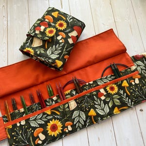 May include: A dark green knitting needle case and matching needle organiser with an orange interior. The exterior features a pattern of sunflowers, mushrooms, and leaves. The case is open, displaying knitting needles and accessories.