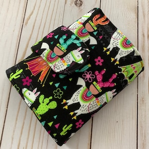May include: Black fabric wallet with a colorful llama print. The llamas are wearing colorful hats and have cacti on their backs. The wallet has a flap closure.