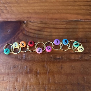 May include: A colorful counting bracelet featuring gold-toned rings and numbered beads. The beads are in various colors, including green, orange, red, pink, and purple, with numbers 0 through 8. The bracelet is designed for educational purposes.