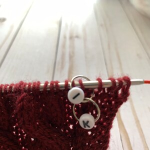 May include: Close-up of knitting needles with a burgundy knitted fabric. Two white stitch markers with the letters "I" and "K" are attached to the needles. The fabric has a textured, patterned design.