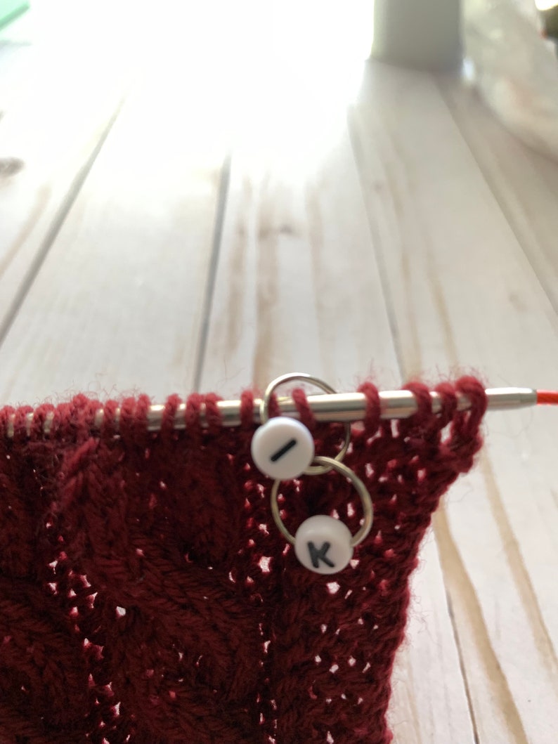 May include: Close-up of knitting needles with a burgundy knitted fabric. Two white stitch markers with the letters "I" and "K" are attached to the needles. The fabric has a textured, patterned design.