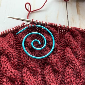 May include: Close-up of a knitting project in progress. The project features a deep red cable knit pattern, with a bright blue spiral stitch marker. Knitting needles are inserted into the stitches, and a ball of red yarn is visible.