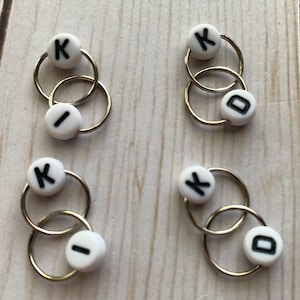 May include: Four sets of interlocking metal rings with white beads. Each bead has a black letter: K, I, or D. The rings are silver-coloured and appear to be jewellery findings or components for crafts.
