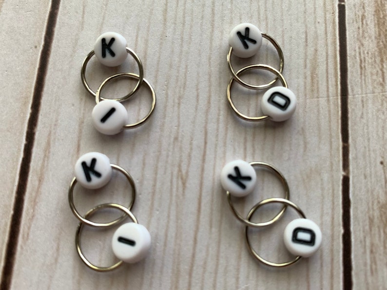 May include: Four sets of interlocking metal rings with white beads. Each bead has a black letter: K, I, or D. The rings are silver-colored and appear to be jewelry findings or components for crafts.