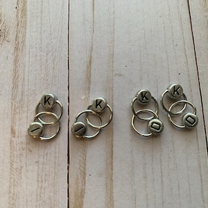 May include: Five silver ring stitch markers with the letter "K" on each. The rings are double-looped and are used for marking rows in knitting.