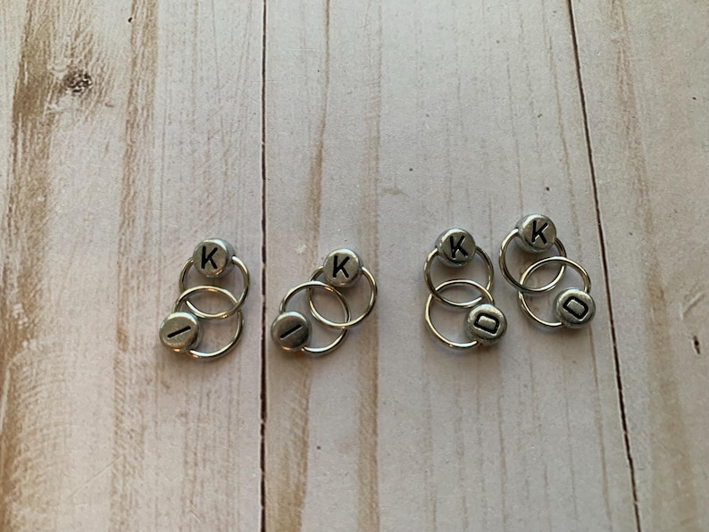 May include: Five silver ring stitch markers with the letter "K" on each. The rings are double-looped and are used for marking rows in knitting.