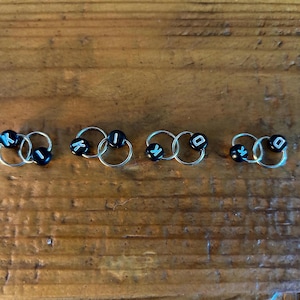 May include: Five pairs of silver hoop earrings, each with two connected hoops. Each pair has black beads with white letters spelling "KIKO" in sequence. The earrings are arranged on a wooden surface.