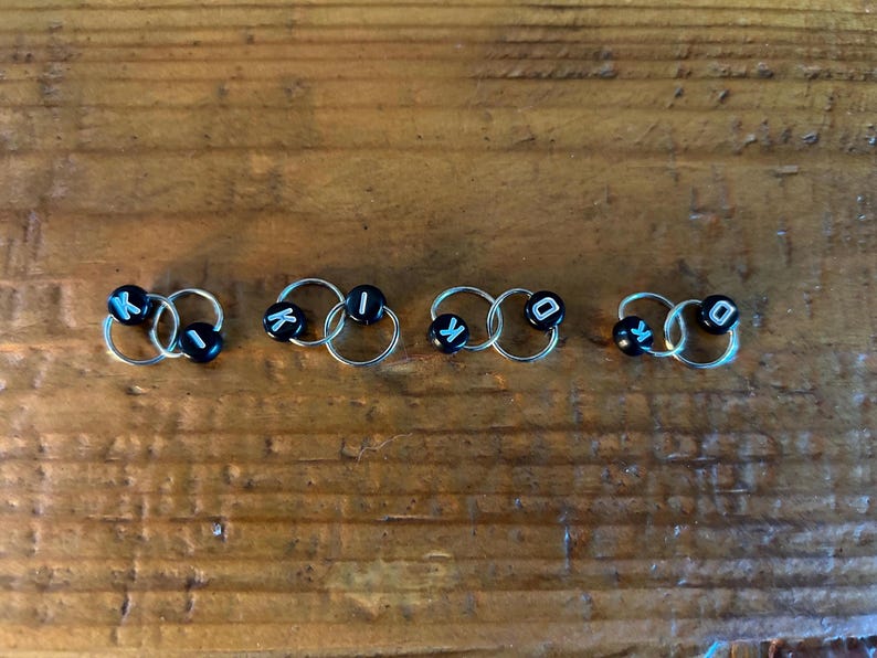 May include: Five pairs of silver hoop earrings, each with two connected hoops. Each pair has black beads with white letters spelling "KIKO" in sequence. The earrings are arranged on a wooden surface.