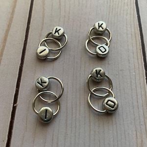 May include: Four sets of silver metal stitch markers with letter beads. Each set has two rings with a bead on each ring. The beads are silver and have the letter "K" on one bead and the letter "I" on the other bead.