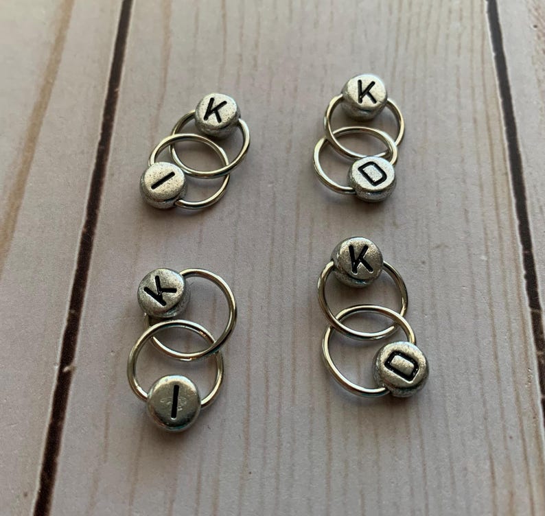 May include: Four sets of silver metal stitch markers with letter beads. Each set has two rings with a bead on each ring. The beads are silver and have the letter "K" on one bead and the letter "I" on the other bead.