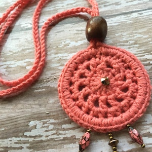 May include: A coral crocheted necklace with a wooden bead and dangling beads. The necklace is on a white wooden surface.