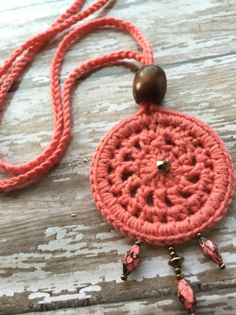 May include: A coral crocheted necklace with a wooden bead and dangling beads. The necklace is on a white wooden surface.