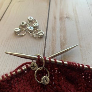 May include: Close-up of a red knitted fabric with two silver knitting stitch markers on a pair of silver knitting needles. The stitch markers are round and have the letters "K" and "I" on them.