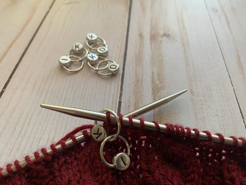 May include: Close-up of a red knitted fabric with two silver knitting stitch markers on a pair of silver knitting needles. The stitch markers are round and have the letters "K" and "I" on them.
