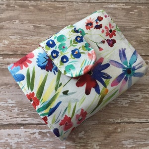 May include: A small white fabric wallet with a floral print featuring blue, red, pink, and green flowers. The wallet has a flap closure.