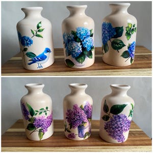May include: Six ceramic vases with floral designs. Three vases feature blue hydrangeas and one has a bluebird. The other three vases feature purple lilac flowers.