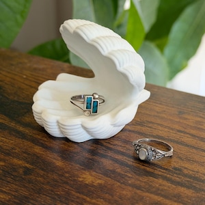 May include: A white seashell-shaped ring holder with two rings. One ring has turquoise-colored rectangular stones, and the other has a white stone. The holder sits on a dark wooden surface.