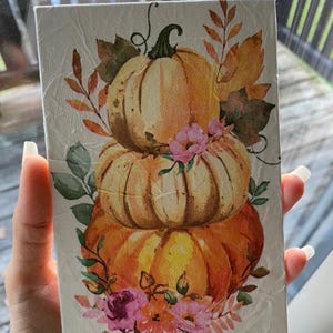 May include: A stack of three pumpkins with orange, yellow, and white paint, decorated with pink and green flowers. The pumpkins are on a white background.