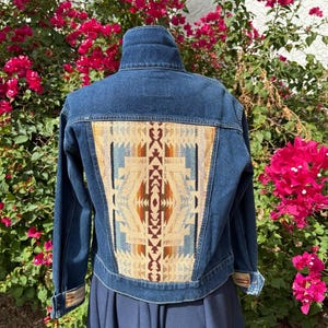 May include: A blue denim jacket with a colourful geometric pattern on the back. The pattern features beige, brown, and blue hues. The jacket has a high collar and long sleeves. The jacket is paired with a navy blue skirt.