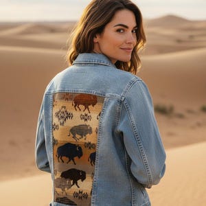 Może przedstawiać: Jasnoniebieska kurtka dżinsowa z ozdobnym panelem z ilustracjami żubrów w odcieniach brązu, beżu i czerni. Kurtka ma klasyczny krój z zapięciami na guziki i kieszeniami. Modelka ma na sobie kurtkę w scenerii pustynnej.