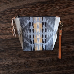 Peut inclure: Une pochette rectangulaire avec un motif géométrique gris, noir et jaune. La pochette a une fermeture éclair noire, une dragonne en cuir beige et une tirette en cuir beige. La pochette est en tissu tissé.
