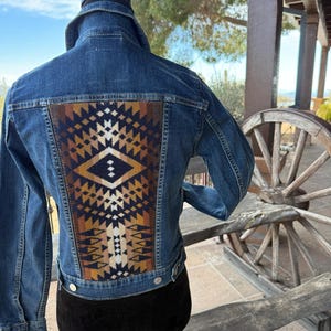 Può includere: Giacca di jeans blu con pannello posteriore a fantasia geometrica. Il pannello presenta un design nei toni del marrone, beige e bianco. La giacca ha un colletto classico, chiusure con bottoni e maniche lunghe. Un capo di abbigliamento elegante.