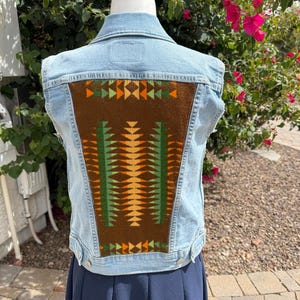 May include: Light blue denim vest with a brown, green, and orange geometric design on the back. The vest has a classic collar, button closures, and a sleeveless design. The back panel features a detailed, patterned fabric.