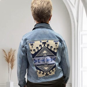 May include: Light blue denim jacket with a geometric design on the back. The design features shades of blue, cream, and black. The jacket has a classic collar and button closures. A stylish outerwear piece.