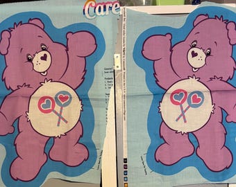 Op de afbeelding: Twee stoffen panelen met een paars Care Bear-personage met een hartvormige neus en een buikbadge met twee hartlolly's. De beer is blauw omlijnd tegen een lichtblauwe achtergrond. Het woord "Care" is bovenaan zichtbaar.