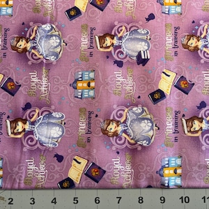 Peut inclure: Tissu violet avec un motif répété d'une princesse en robe bleue, un livre, un bâtiment et le texte "Princess in training" et "Royal Achiever".