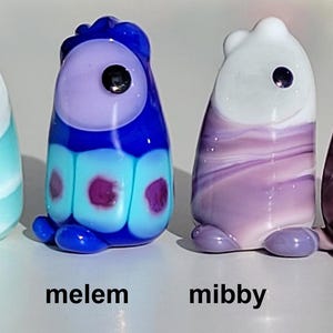 May include: Four colorful glass figurines in a row, each with a unique design and color scheme. The figurines are named "marty", "melem", "mibby", and "mauvlus". "Marty" is light blue, "melem" is blue and purple, "mibby" is purple, and "mauvlus" is pink with a flower.