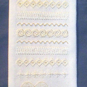 Op de afbeelding: Witte stoffen banner met geborduurde ontwerpen, waaronder twee vogels, harten en verschillende steekpatronen. De banner hangt aan een gouden hartvormige hanger en heeft een gouden kwast.