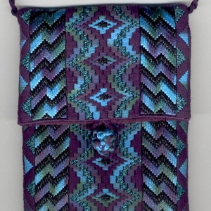 Peut inclure: Un petit sac en tissu violet avec un motif géométrique bleu et vert. Le sac a une bandoulière en cordon tressé et une fermeture à glands.