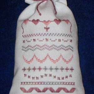 May include: A white fabric drawstring bag with pink and gray embroidery. The embroidery features a variety of patterns, including hearts, stars, and waves.