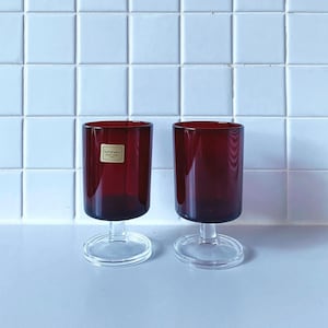 Op de afbeelding: Twee heldere glazen wijnglazen met een rode basis. De glazen hebben een kleine steel en een ronde voet. Het glas links heeft een klein label met de tekst "lumina-c".