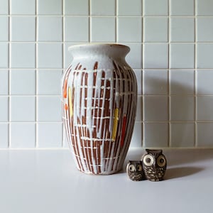 Könnte beinhalten: Eine weiße Keramikvase mit braunen und orangefarbenen geometrischen Mustern. Zwei kleine braune Keramik-Eulenfiguren stehen vor der Vase.