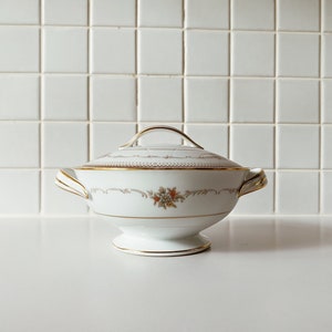 Puede incluir: Azucarero de porcelana blanca con ribete dorado y diseño floral. El tazón tiene una tapa y dos asas.