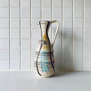 Może przedstawiać: Wazon ceramiczny w stylu vintage z kremową podstawą, ozdobiony czarnymi pionowymi liniami i kolorowymi abstrakcyjnymi kształtami w kolorze żółtym, niebieskim i czerwonym. Dzbanek ma zakrzywiony uchwyt i wylewkę.