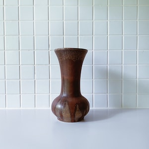Könnte beinhalten: Eine braune Keramikvase mit einer strukturierten Oberfläche und einem goldfarbenen Design. Die Vase hat einen schmalen Hals und einen runden Körper.