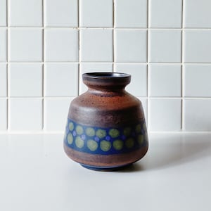 May include: A ceramic vase with a brown and blue color scheme. The vase has a rounded body with a narrow neck and a dark rim. A blue band with green and purple dots encircles the middle of the vase. The vase is likely used for decorative purposes.
