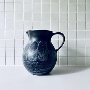 May include: A dark blue ceramic pitcher with a rounded body and a curved handle. The surface features a textured, mottled glaze pattern with lighter blue and grey arched designs around the upper section and horizontal bands near the base.